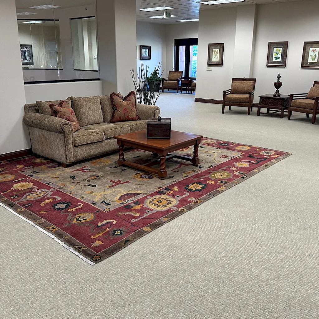 Commercial lobby with beige carpet, ornate red and gold area rug, sofa seating, and floral arrangements.