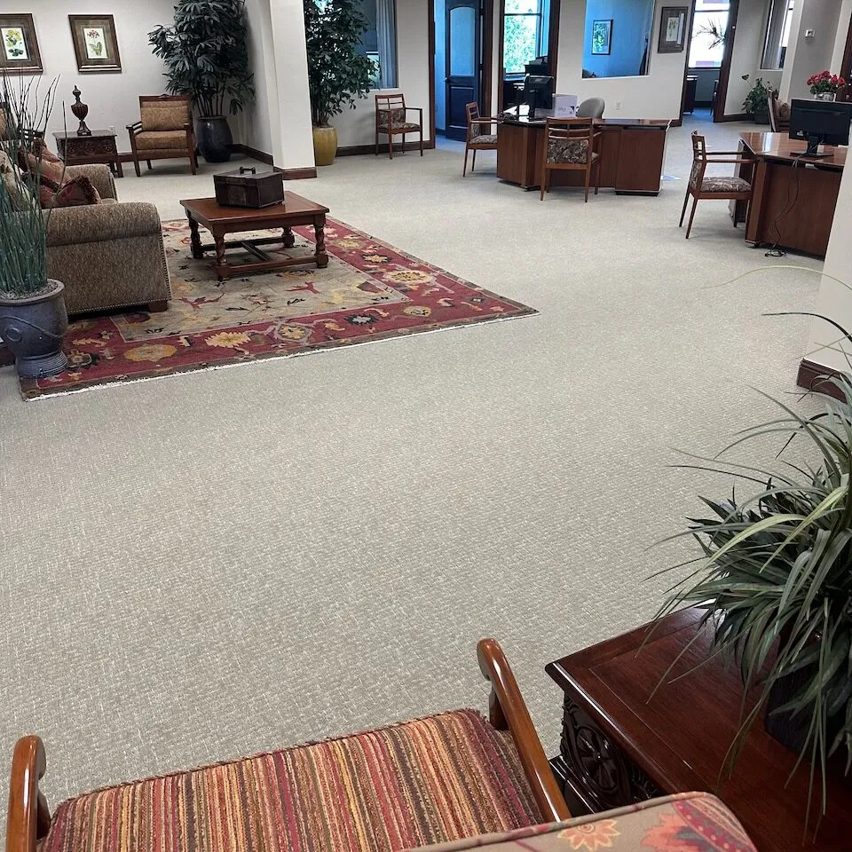 Large commercial office lobby with beige carpet, decorative area rug, desks, and indoor plants.