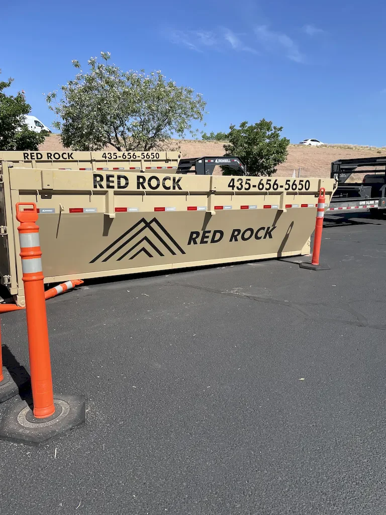 Tan Red Rock branded roll-off dumpster on asphalt with orange safety delineators and mountain logo on the side.