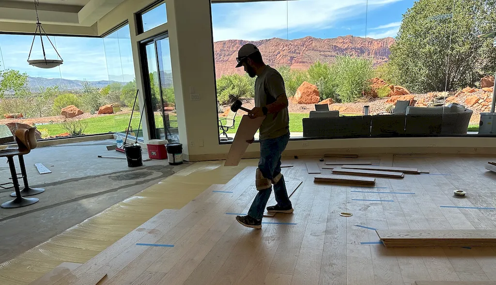 Red Rock Flooring installer laying light wood planks with knee pads and mallet before Southern Utah red rock mountain windows.