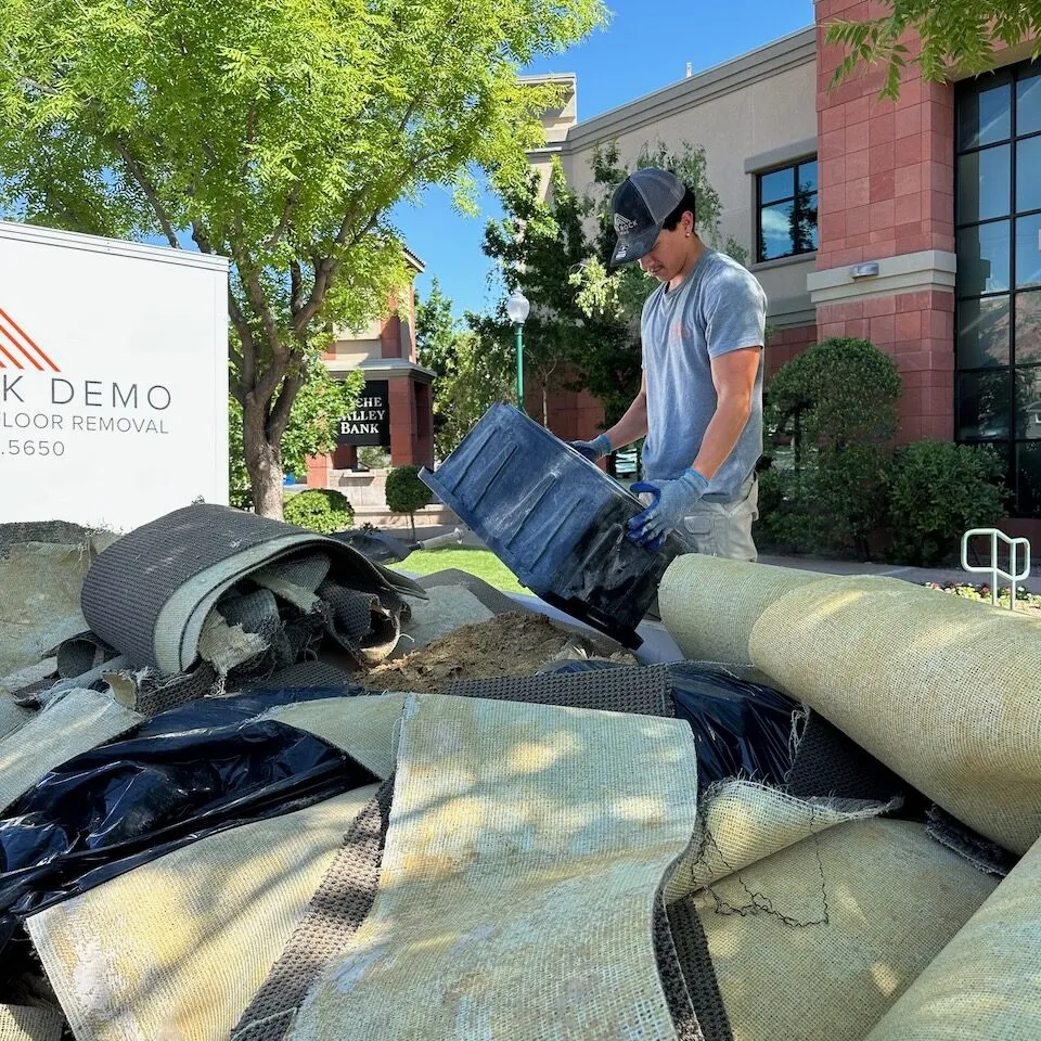 Red Rock Flooring worker emptying a debris bin beside a company demo floor removal truck loaded with carpet scraps.