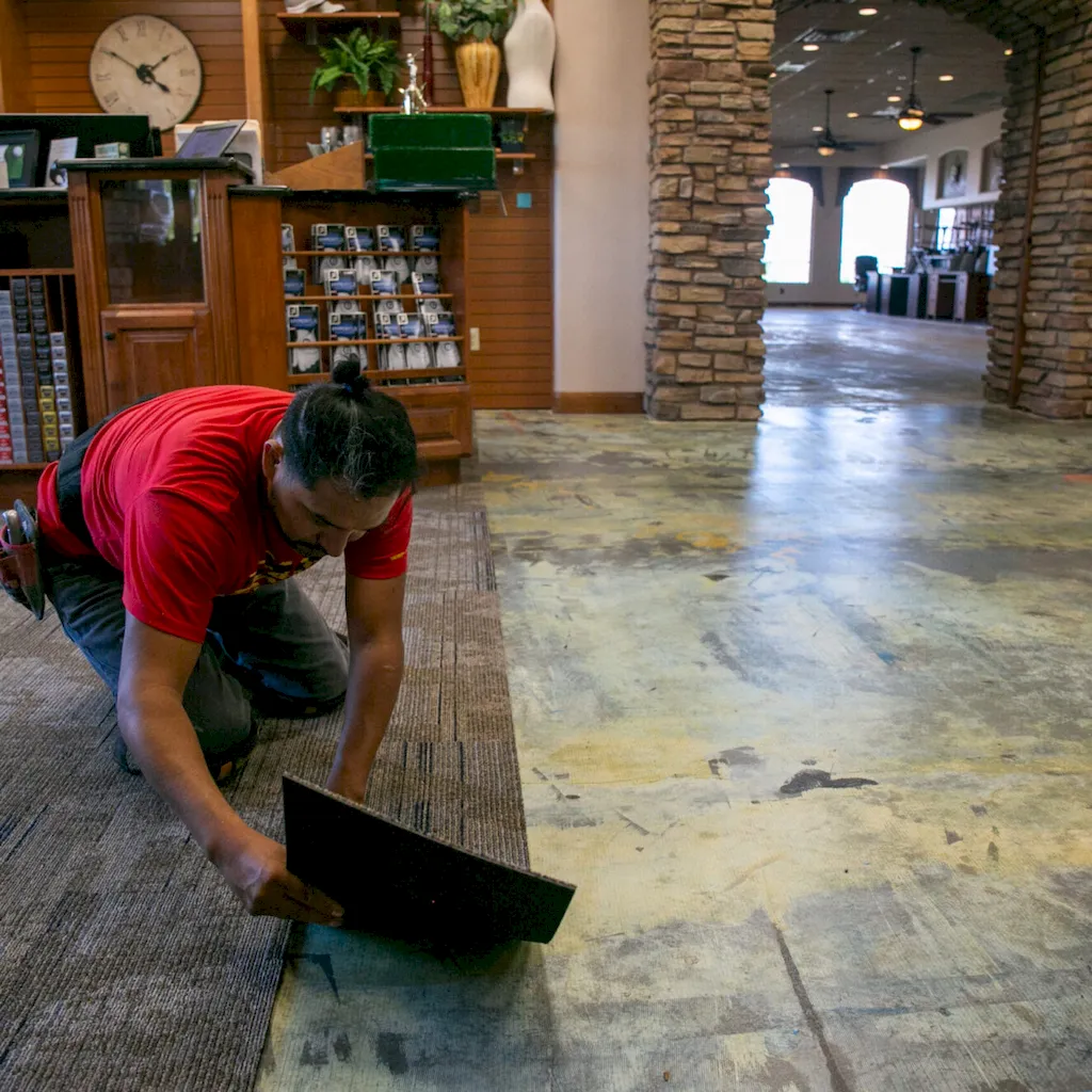 Red Rock Flooring worker in a red shirt kneeling to install brown carpet tiles in a commercial retail space.